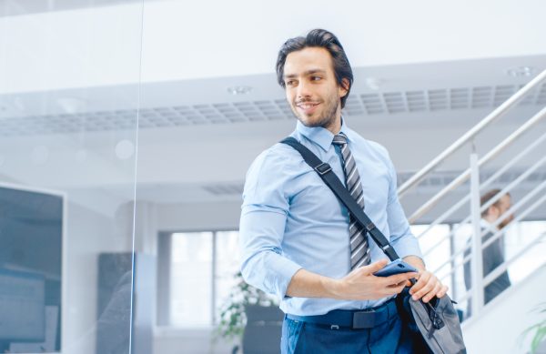 Young Happy Businessman Walks Into Bright Modern Office, Takes Place at His Desktop Computer. In the Background Diverse Team of Professional Businesspeople Working
