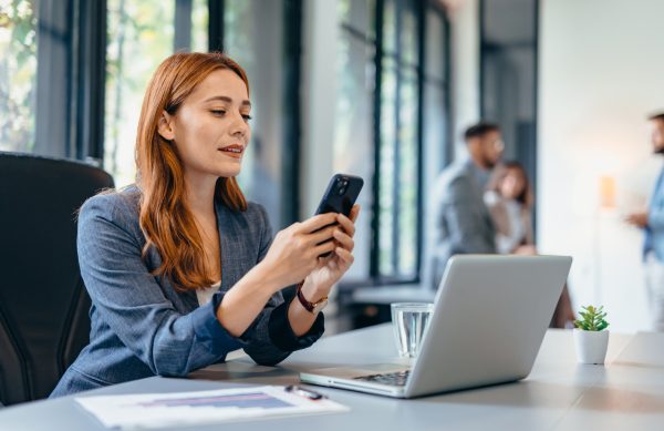 A professional woman interact with her smartphone while working in a modern well-lit office. In the background other employees can be seen collaborating.